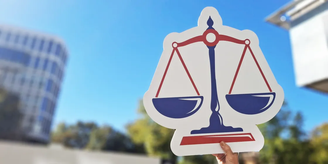 Das Schild einer Wage wird von einer Hand in den Himmel gehalten. Das Schild zeigt eine Waage, die für die Rechtsprechung steht.