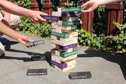 Hände, die einen Jenga-Turm bauen, dessen Steine mit Schlüsselwörtern aus der Bildungsarbeit versehen sind